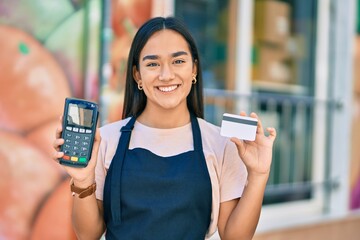 Young latin shopkeeper girl smiling happy holding credit card and dataphone at fruit store.