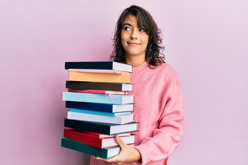 Young hispanic woman holding a pile of books smiling looking to the side and staring away thinking.