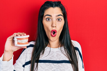 Young hispanic girl holding denture scared and amazed with open mouth for surprise, disbelief face