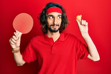 Young hispanic man holding red ping pong racket and ball smiling looking to the side and staring away thinking.