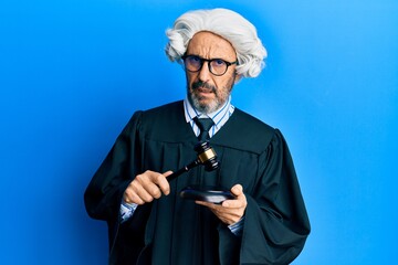 Middle age hispanic man using gavel in shock face, looking skeptical and sarcastic, surprised with...