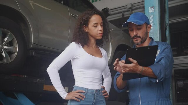 Young mechanic stand and talking to customer about car with repairing in the garage, auto service, technician man explaining problem of vehicle and look clipboard with client, transportation concept.