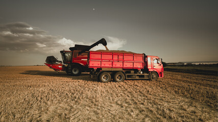 Harvesting grain in a farmer's field. The combine harvester unloads into the back of an agricultural the truck in the evening at sunset. Bright rural background, wallpaper, design. Tinted photo.