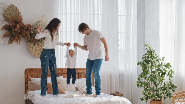 Young parents married couple caucasian family with little daughter toddler child girl jumping on bed holding hands jump into air in bedroom having fun playing at home enjoy parenting and communication