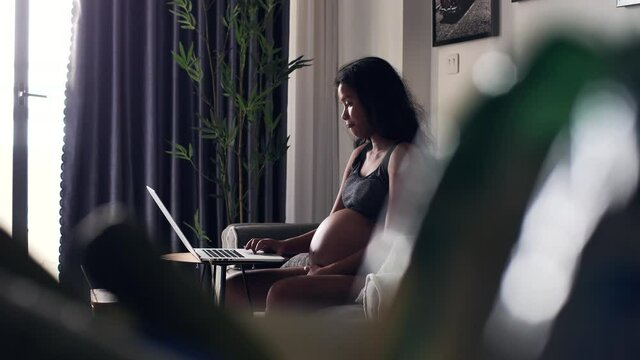 Young pregnant woman gently touching her tummy while working online on laptop.