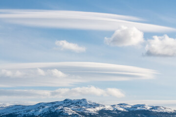 Wispy, unique and strange looking clouds with blue sky background and snow capped mountains below. 