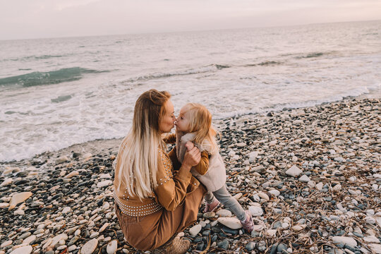 Mother with baby daughter next to the sea smiling. Baby girl with blonde hair, in a warm fur vest. Spring time, sun is shining. Travel abroad. Happy young family. 