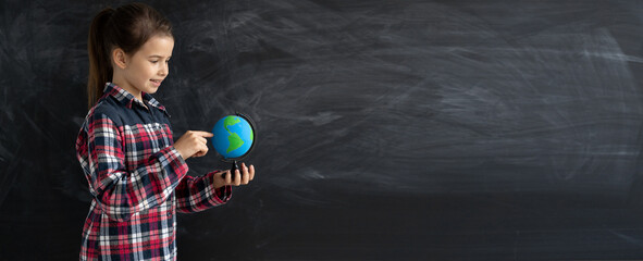 young caucasian girl schoolgirl or student stands near the chalk blackboard. Holds a globe. Baner
