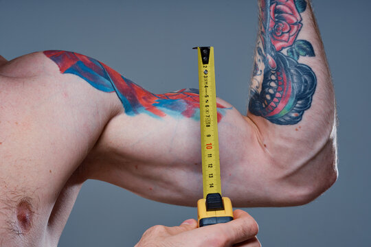 guy measures arm muscles with centimeters on a gray background and a multicolored tattoo bodybuilder fitness