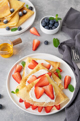 Crepes or thin pancakes with cream cheese, fresh strawberries and honey on a white plate on a gray concrete background. Copy space.
