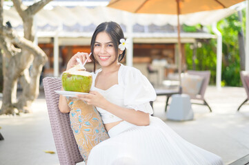 Beautiful woman tourist with white flower on her hair drinking coconut sitting on lounge chair during summer holidays