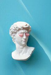 Plaster head of David with pink hearts on eyes, blue background. White sculpture of antique head in...