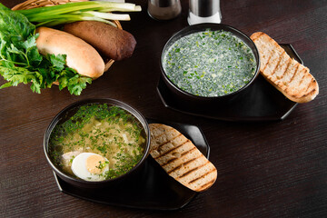 Okroshka, broth with noodles and eggs in black plate, toasted bread, herbs on dark wooden table....