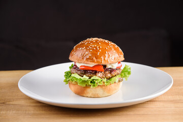 Burger with cutlet, sauce, tomato, lettuce, sesame on white plate on dark background.