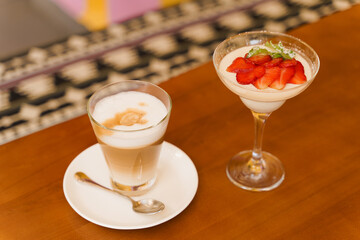 Creamy dessert with strawberries in glass, cappuccino on wooden table. Appetizing dessert and latte on the background of the restaurant
