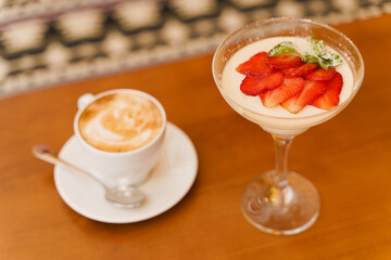Creamy dessert with strawberries in glass, cappuccino on wooden table. Appetizing dessert and latte on the background of the restaurant
