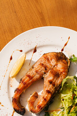 Grilled salmon steak with lettuce and lemon on white plate on wooden table