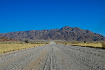 Fototapeta na wymiar Gravel road leading towards mountains in the Namibian desert on a sunny day with blue sky and no clouds