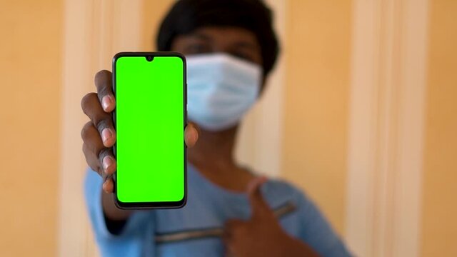 Portrait of an Indian kid wearing face mask showing mobile phone with green screen towards the camera and giving thumbs up
