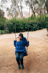 Mom in a blue warm sweater and jeans is swinging on a swing with a tiny baby on her chest