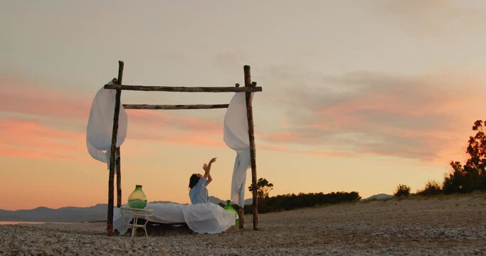 Giovane ragazza osserva il tramonto in un letto a baldacchino sulla spiaggia in riva al mare