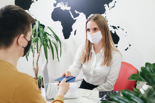 Travel agent having a client in face mask in modern office of tour agency, giving passport after check-in or booking flight. Assistant keeps tickets for the plane in the travel agency to clients. 