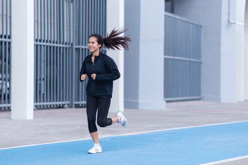 Asian Beautiful Young woman black suit with happy running or jogging on blue running track in the day time. exercise, training, fitness, workout, sport, lifestyle and healthy concept.