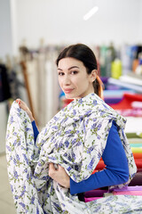 Tailoring, seamstress or atelier concept. Beautiful brunette fabrics store customer or buyer matching textile linen cloth on herself, applying it to the body in front of the mirror. High quality image