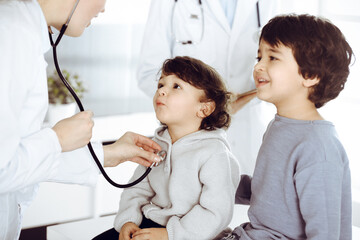 Fototapeta premium Woman-doctor examining a child patient by stethoscope. Cute arab toddler and his brother at physician appointment. Medicine concept