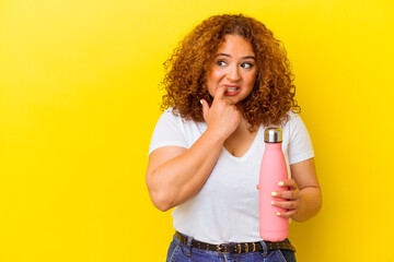 Young latin woman holding a thermos isolated on yellow background relaxed thinking about something looking at a copy space.