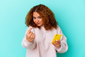 Young latin woman holding mobile phone isolated on blue background pointing with finger at you as if inviting come closer.