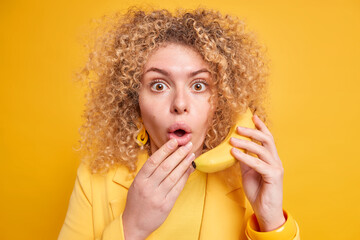 Impressed woman with healthy skin curly bushy hair keeps mouth opened from amazement holds ripe banana near ear pretends telephone conversation wears yellow clothes in one tone with background
