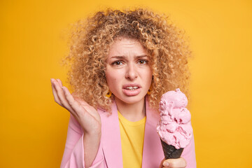 Frustrated displeased woman with curly hair raises palm looks disappointed dressed formally holds appetizing cold ice cream in black waffle doesnt like something isolated over yellow studio wall
