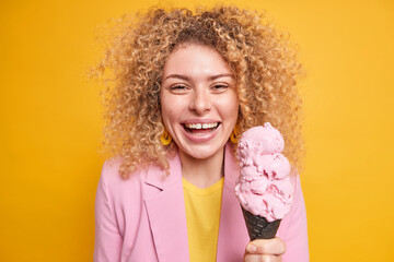 Portrait of pleasant looking woman with curly hair holds cone ice cream has day off walks with friends in park during summer time isolated over yellow background. Female model eats yummy dessert