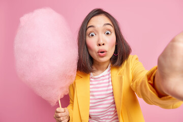 Studio shot of startled Asian woman stares bugged eyes at camera makes photo of herslef holds tasty cotton candy likes eating something sweet poses against pink background in stylish clothes