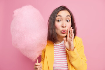 Studio shot of beautiful young woman with dark hair keeps hand near mouth whispers something has surprised expression holds sweet candy floss wears yellow jacket isolated over pink background