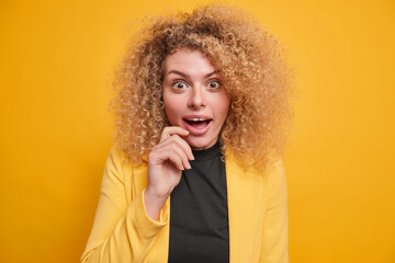 Portrait of surprised curly haired young woman has cheerful happy expression stares shocked at camera cannot believe in something great dressed formally isolated over vivid yellow background.