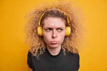 Headshot of displeased curly haired teenage girl has bad mood sulking face expression wears wireless stereo headphones listens music dressed in casual black t shirt isolated over yellow background