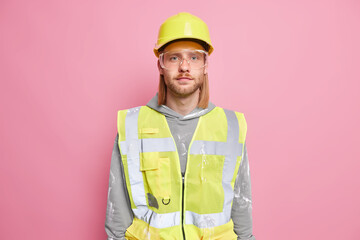 Serious man builder engineer wears construction safety helmet uniform glasses looks confidently at camera ready for work isolated over pink background. Self assured workman or construction worker