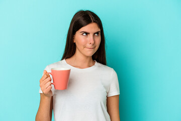 Young caucasian woman holding a mug isolated on blue background confused, feels doubtful and unsure.