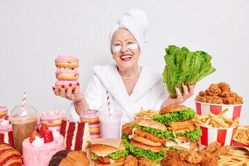 Positive old wrinkled woman smiles happily chooses between green lettuce salad and doughnuts has good mood surrounded by high calorie snacksapplies patches under eyes dressed in soft white bathrobe