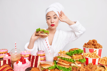 Self confident Asian woman looks attentively at camera holds delicious appetising hamburger wears bathrobe and towel on head poses near variety of junk food has unhealthy ration. Tasty snack