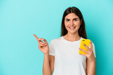 Young caucasian woman holding a mobile phone isolated on blue background smiling and pointing aside, showing something at blank space.