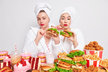 Binge eating habit concept. Two serious women wear red lipstick hold tasty hamburgers wear white soft bath dressing gowns and wrapped towels on head isolated over white background. Fast food eaters