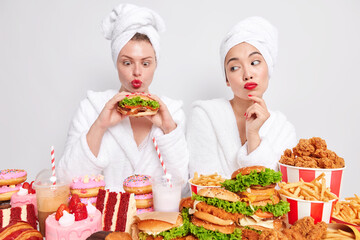 Funny woman holds appetizing delicious burger keeps lips rounded starves to eat delicious snack addicted to cheat meal. Serious Asian lady with red lips weas shower towel and dressing gown temptating