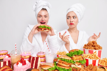 Unhealthy nutrition and food choice concept. Hungry young surprised woman eats delicious sandwich keeps mouth wide opened her friend stand near and looks with temptation at cheat meal keeps to diet