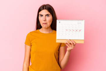 Fototapeta na wymiar Young caucasian woman holding calendar isolated on pink background shrugs shoulders and open eyes confused.