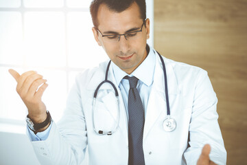 Man-doctor sitting at the desk at his working place and smiling. Perfect medical service in sunny clinic