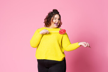 Happy plus size model in casual clothes with big lollipop, fat woman in yellow jumper on pink background