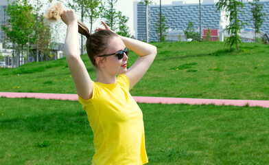 Young woman wearing yellow T-shirt is making a ponytail preparing for a workout or jogging in the city park. Spending time outdoors - obrazy, fototapety, plakaty